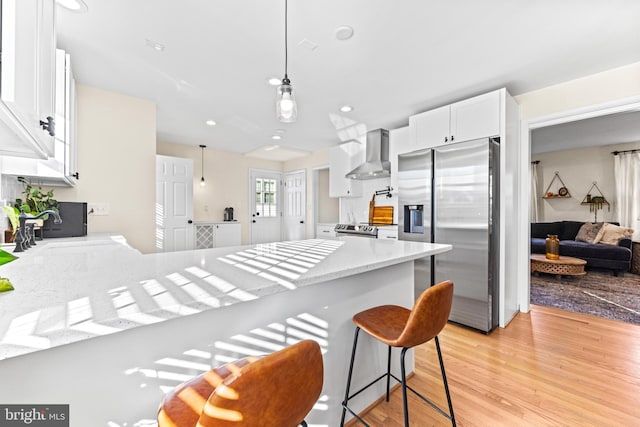 kitchen with stainless steel appliances, wall chimney range hood, decorative light fixtures, light hardwood / wood-style floors, and white cabinetry