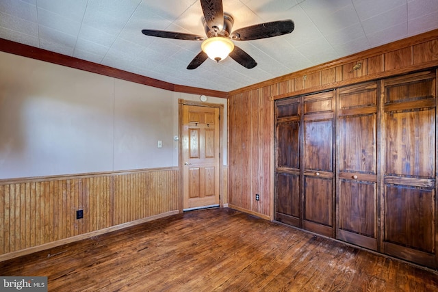 unfurnished bedroom with ceiling fan, dark hardwood / wood-style floors, crown molding, wooden walls, and a closet