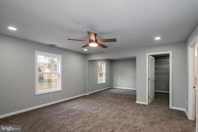 unfurnished bedroom with a walk in closet, ceiling fan, multiple windows, and dark colored carpet