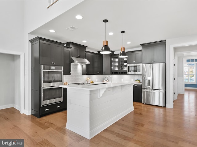 kitchen featuring appliances with stainless steel finishes, light wood-type flooring, backsplash, a center island with sink, and decorative light fixtures