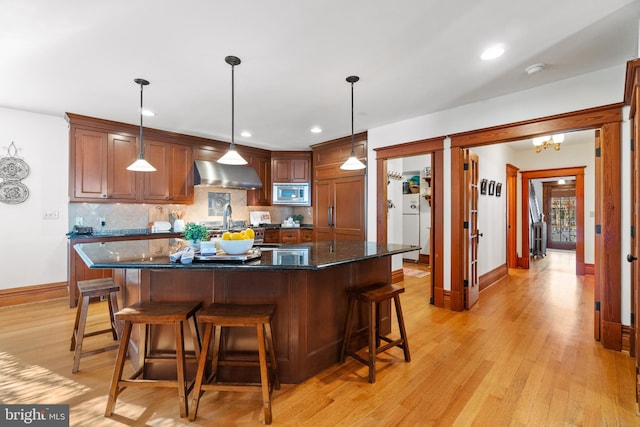 kitchen featuring dark stone counters, wall chimney range hood, pendant lighting, light hardwood / wood-style floors, and stainless steel microwave