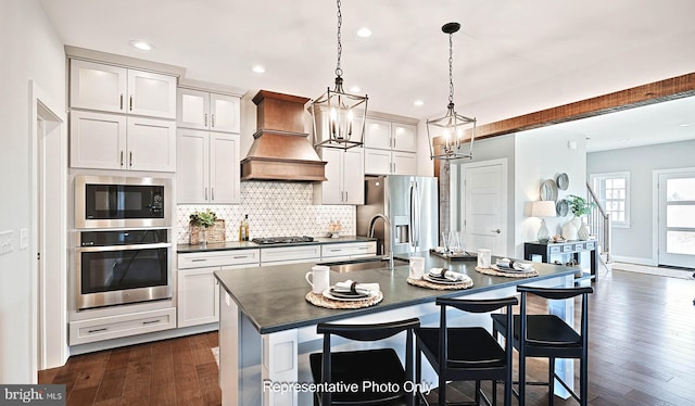 kitchen featuring appliances with stainless steel finishes, premium range hood, dark wood-type flooring, decorative light fixtures, and an island with sink