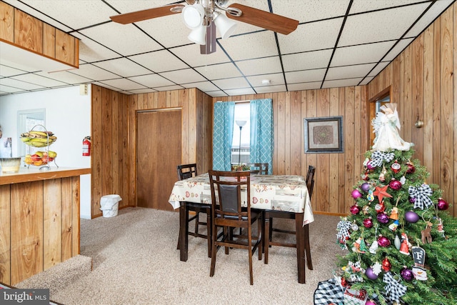 carpeted dining area with a paneled ceiling, wood walls, and ceiling fan