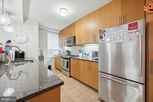 kitchen featuring sink, stainless steel appliances, dark stone counters, decorative light fixtures, and light wood-type flooring