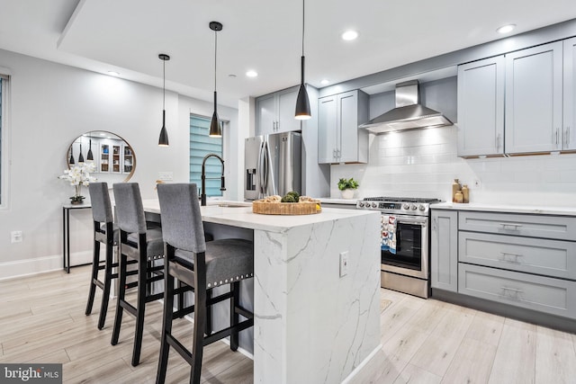 kitchen with gray cabinetry, wall chimney exhaust hood, an island with sink, appliances with stainless steel finishes, and light hardwood / wood-style floors