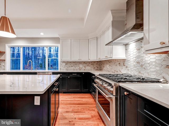 kitchen featuring wall chimney range hood, light hardwood / wood-style flooring, white cabinets, stainless steel stove, and hanging light fixtures