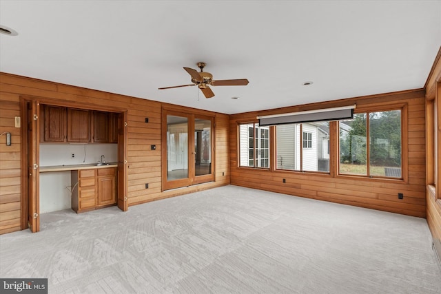 unfurnished living room featuring light carpet, sink, wooden walls, ceiling fan, and built in desk