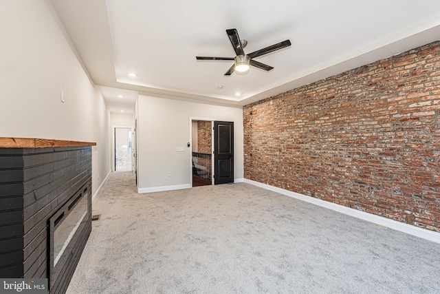 unfurnished living room with light colored carpet, ceiling fan, a tray ceiling, and brick wall