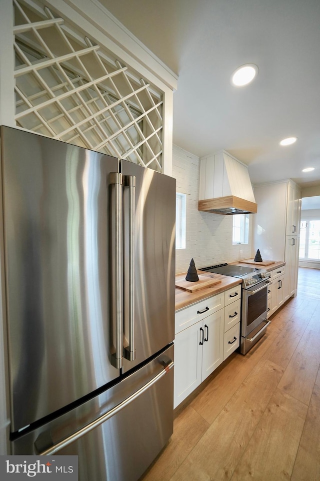 kitchen with wood counters, appliances with stainless steel finishes, custom range hood, white cabinets, and light hardwood / wood-style floors