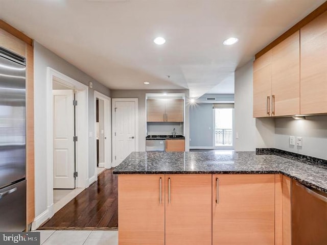 kitchen with kitchen peninsula, light brown cabinetry, light wood-type flooring, appliances with stainless steel finishes, and dark stone countertops