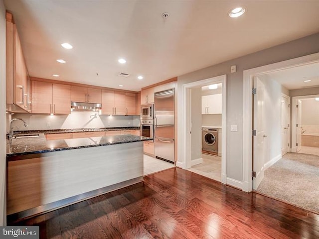 kitchen featuring sink, light hardwood / wood-style flooring, built in appliances, dark stone countertops, and washer / clothes dryer