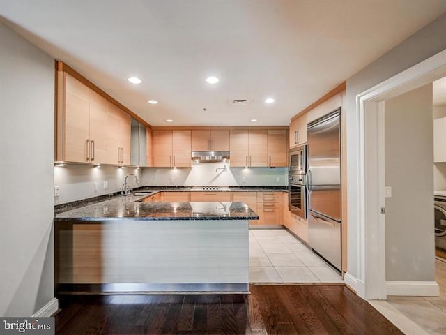 kitchen featuring kitchen peninsula, dark stone counters, light brown cabinets, built in appliances, and light hardwood / wood-style floors