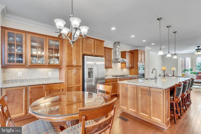 kitchen featuring hanging light fixtures, stainless steel appliances, wall chimney range hood, light stone counters, and light hardwood / wood-style flooring