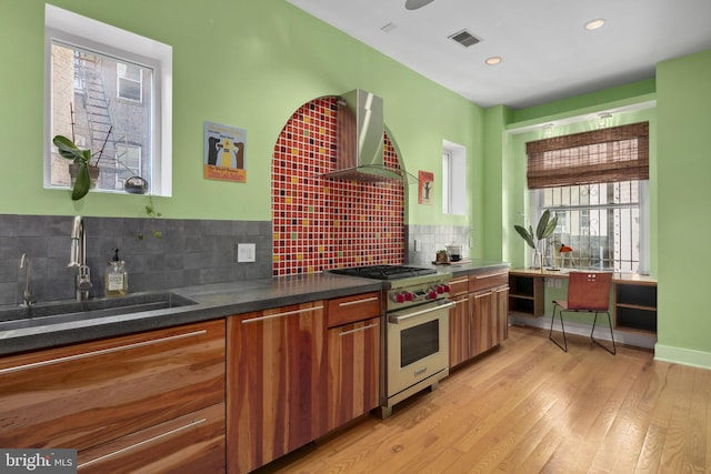 kitchen featuring sink, wall chimney exhaust hood, decorative backsplash, designer range, and light wood-type flooring