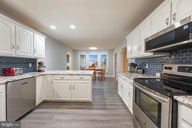 kitchen with light stone countertops, appliances with stainless steel finishes, kitchen peninsula, hardwood / wood-style flooring, and white cabinets