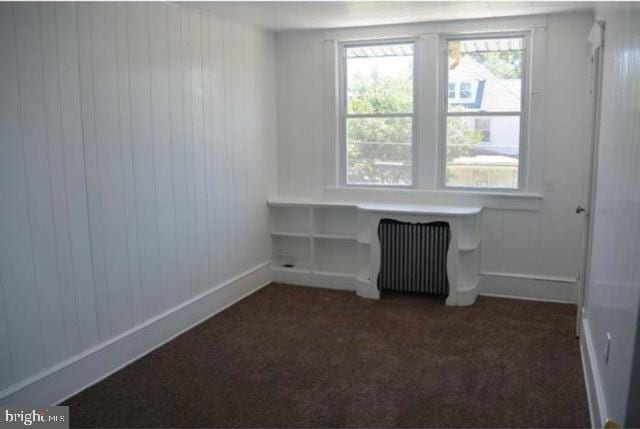 empty room featuring dark colored carpet, wooden walls, and radiator