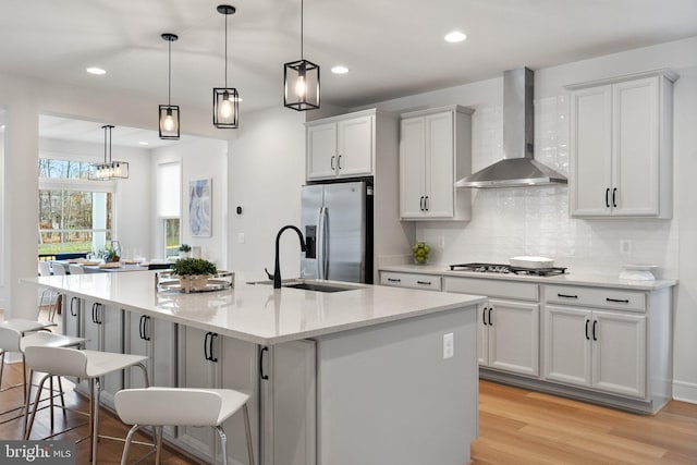 kitchen with wall chimney range hood, an island with sink, light hardwood / wood-style floors, a breakfast bar, and appliances with stainless steel finishes