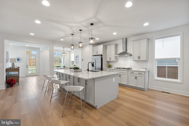 kitchen with stainless steel appliances, a kitchen island with sink, wall chimney range hood, white cabinetry, and hanging light fixtures