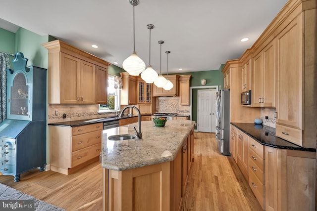 kitchen with dark stone countertops, decorative light fixtures, a kitchen island with sink, appliances with stainless steel finishes, and light wood-type flooring