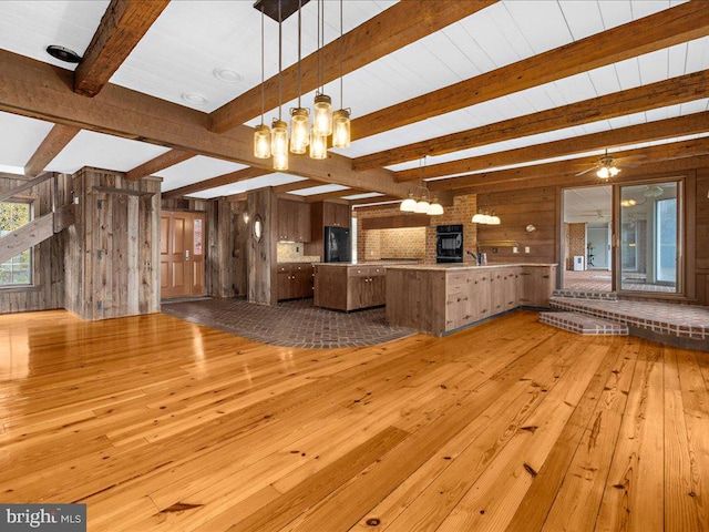 unfurnished living room featuring beam ceiling, sink, wood walls, ceiling fan with notable chandelier, and light wood-type flooring