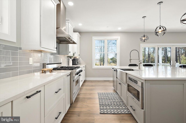 kitchen with white cabinets, wall chimney exhaust hood, an island with sink, light hardwood / wood-style floors, and stainless steel appliances