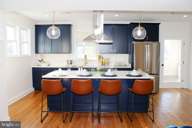 kitchen with stainless steel refrigerator, blue cabinets, island exhaust hood, a kitchen island with sink, and light wood-type flooring