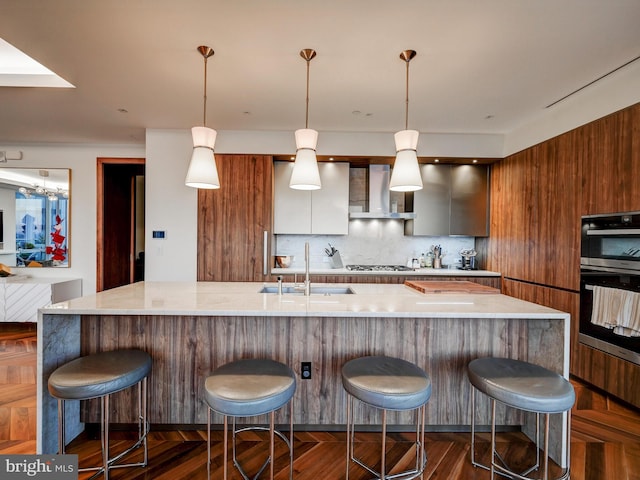 kitchen featuring dark wood-type flooring, a kitchen breakfast bar, wall chimney range hood, sink, and decorative light fixtures