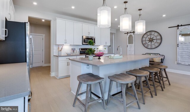 kitchen with white cabinetry, hanging light fixtures, a barn door, an island with sink, and appliances with stainless steel finishes