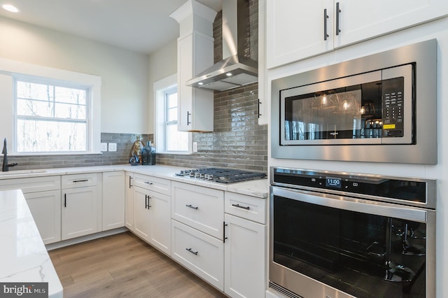 kitchen with white cabinetry, appliances with stainless steel finishes, wall chimney range hood, and light stone counters