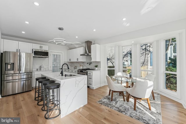 kitchen featuring appliances with stainless steel finishes, wall chimney range hood, white cabinets, light hardwood / wood-style floors, and an island with sink