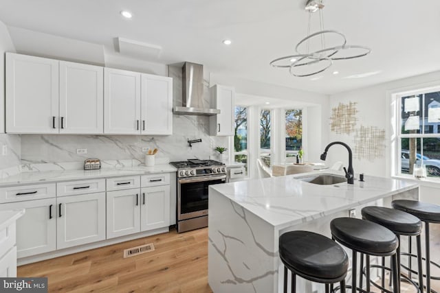 kitchen with gas range, a kitchen island with sink, wall chimney range hood, white cabinets, and hanging light fixtures