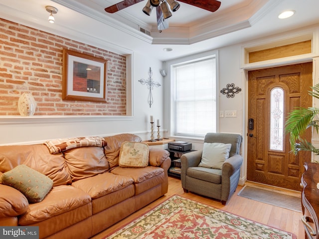 living room with ceiling fan, brick wall, crown molding, a tray ceiling, and light wood-type flooring