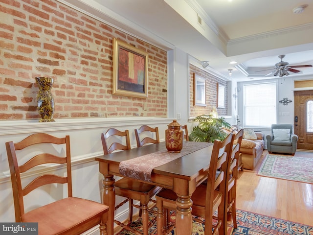 dining space featuring a raised ceiling, ceiling fan, ornamental molding, wood-type flooring, and brick wall