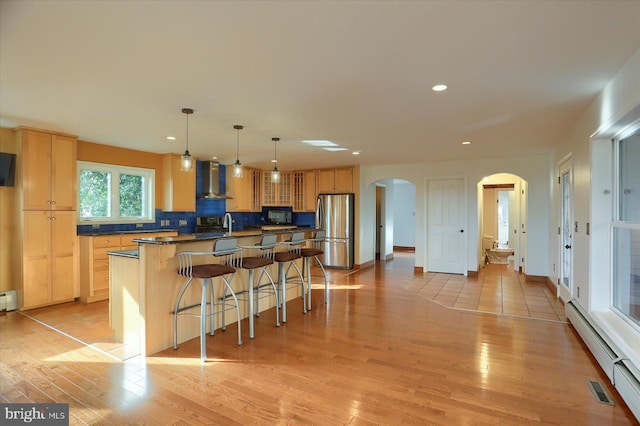 kitchen with stainless steel fridge, baseboard heating, wall chimney range hood, decorative light fixtures, and light hardwood / wood-style floors