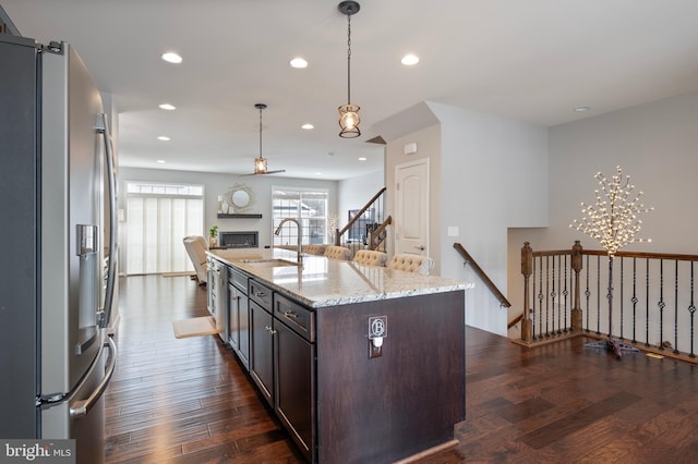 kitchen with decorative light fixtures, sink, stainless steel fridge, a kitchen island with sink, and light stone counters