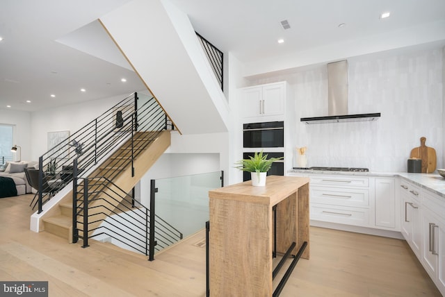 kitchen featuring white cabinetry, wall chimney exhaust hood, stainless steel gas cooktop, double oven, and light wood-type flooring