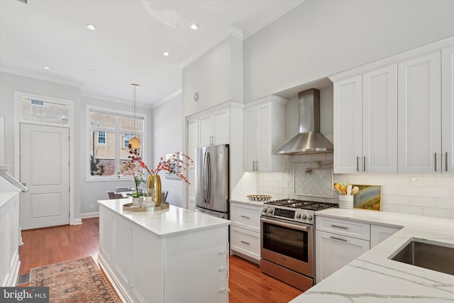kitchen with ornamental molding, wall chimney exhaust hood, white cabinets, appliances with stainless steel finishes, and backsplash