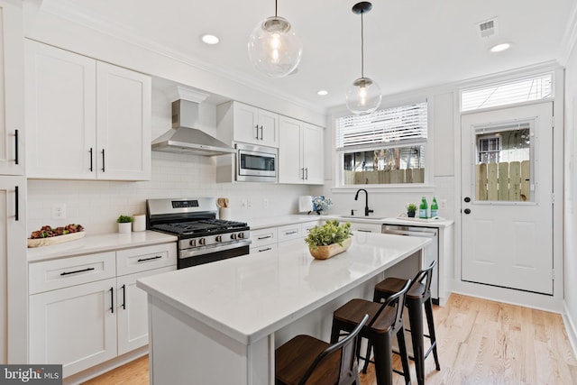 kitchen featuring stainless steel appliances, wall chimney range hood, decorative light fixtures, a center island, and white cabinetry