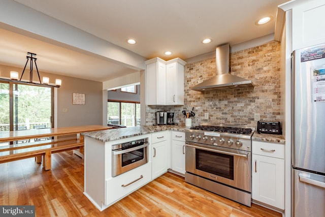 kitchen featuring kitchen peninsula, appliances with stainless steel finishes, wall chimney exhaust hood, light hardwood / wood-style flooring, and white cabinetry