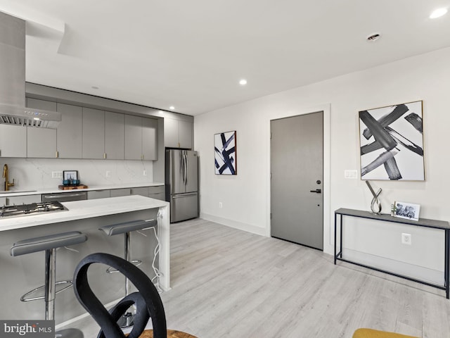 kitchen with gray cabinets, light wood-type flooring, range hood, appliances with stainless steel finishes, and a breakfast bar area