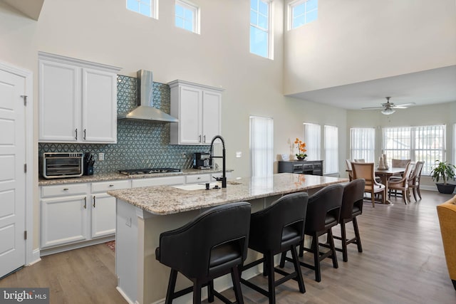 kitchen featuring a wealth of natural light, a center island with sink, a high ceiling, and wall chimney range hood