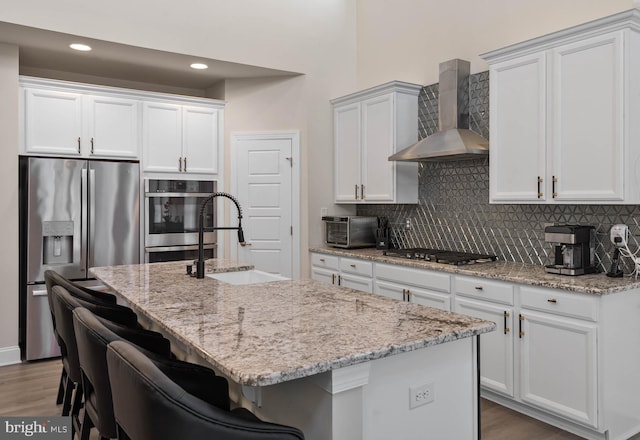 kitchen with white cabinets, hardwood / wood-style flooring, wall chimney exhaust hood, an island with sink, and stainless steel appliances