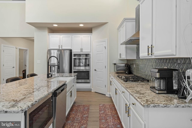 kitchen with light stone countertops, appliances with stainless steel finishes, wall chimney range hood, light hardwood / wood-style floors, and white cabinetry