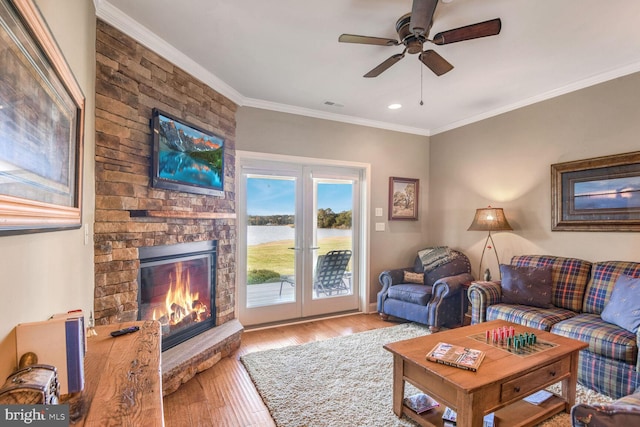 living room featuring ceiling fan, crown molding, a water view, light hardwood / wood-style floors, and a stone fireplace