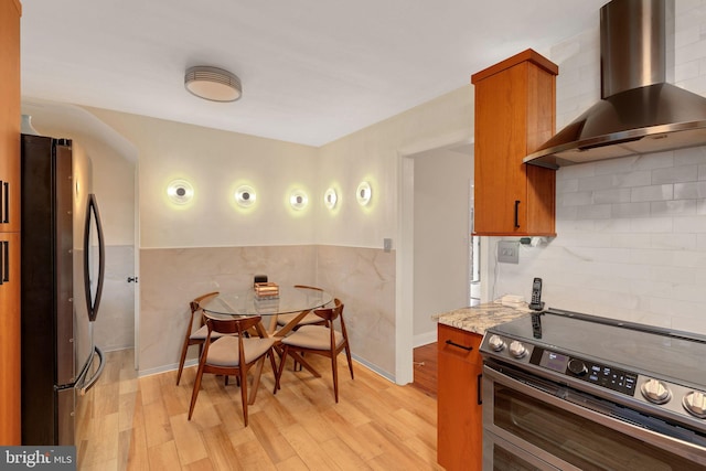 kitchen featuring light wood-type flooring, light stone counters, stainless steel appliances, wall chimney range hood, and tile walls