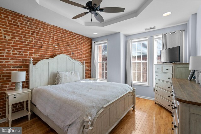 bedroom with a raised ceiling, brick wall, ceiling fan, and light hardwood / wood-style floors