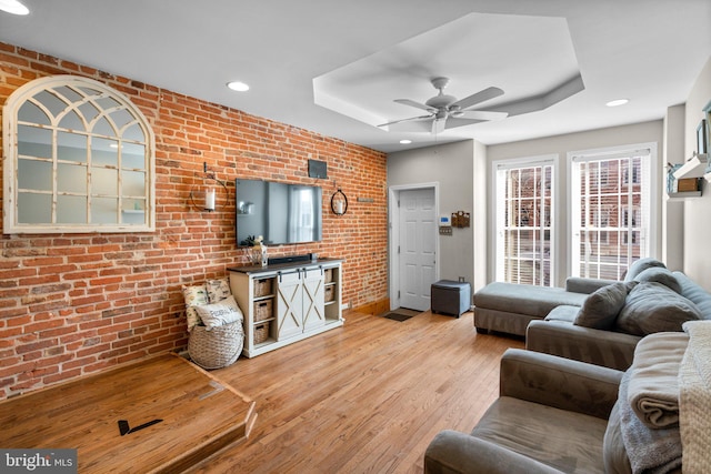 living room with light hardwood / wood-style floors, ceiling fan, a tray ceiling, and brick wall