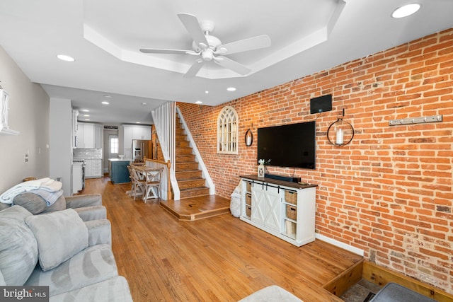 living room with light hardwood / wood-style flooring, a raised ceiling, ceiling fan, and brick wall