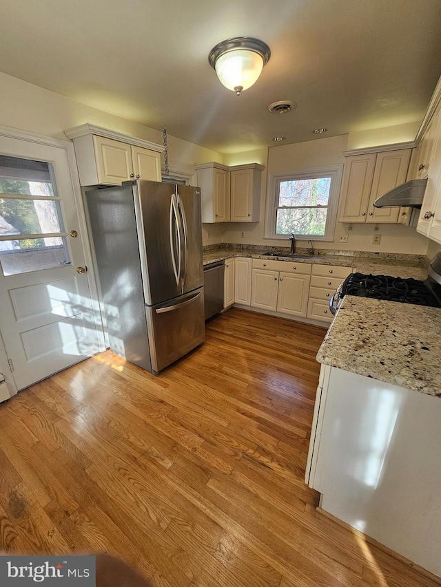 kitchen with light wood-type flooring, range hood, light stone counters, and appliances with stainless steel finishes