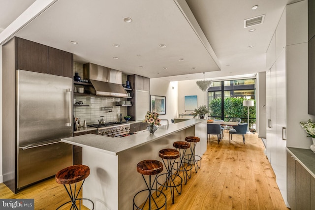 kitchen featuring visible vents, high end appliances, wall chimney exhaust hood, modern cabinets, and a kitchen bar